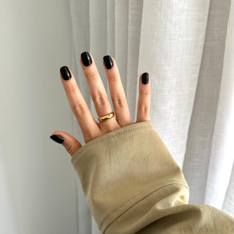 A hand with black press on nails wearing a gold ring on the ring finger, with the rest of the hand covered by a beige sleeve.