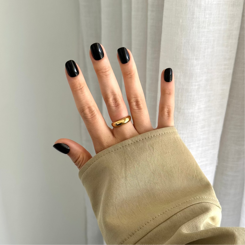 A hand with black press on nails wearing a gold ring on the ring finger, with the rest of the hand covered by a beige sleeve.