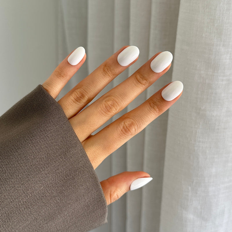 A hand with white oval-shaped press on nails, partially covered by a gray sleeve, against a translucent curtain background.