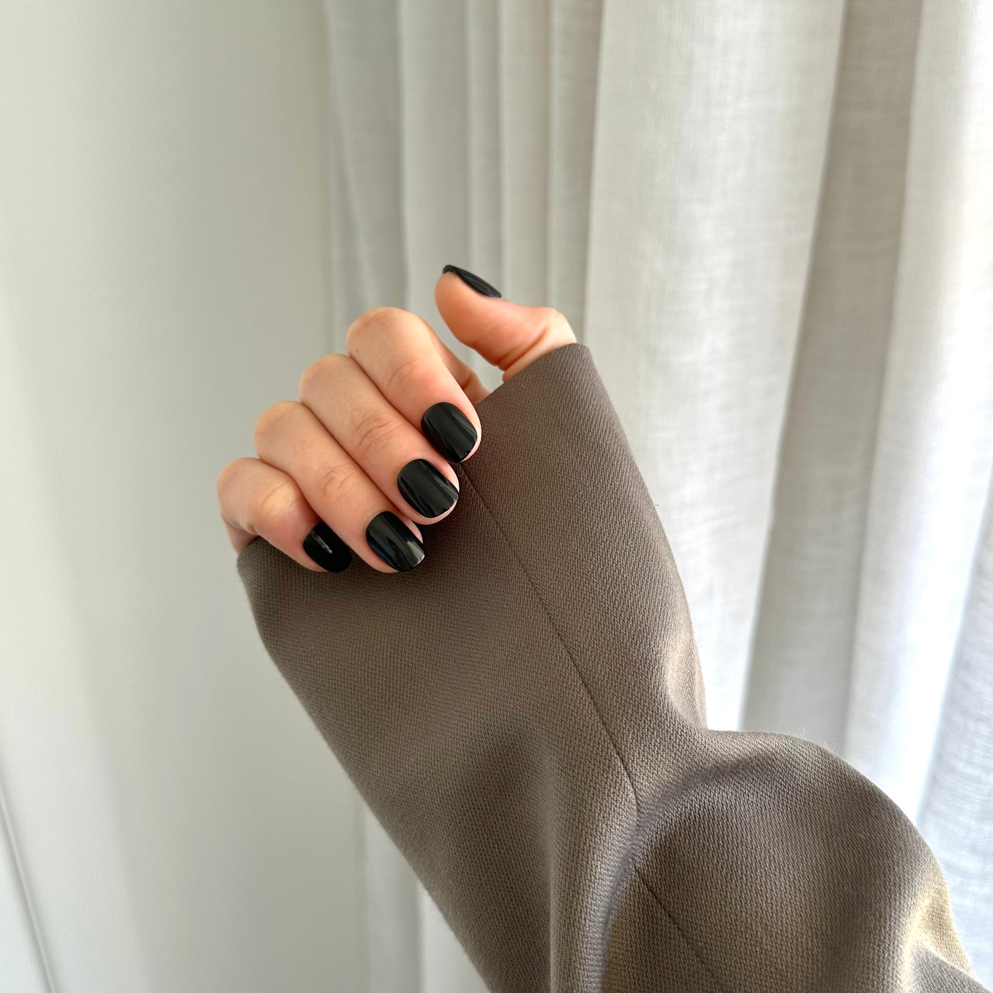 A hand with black press on nails wearing a gold ring on the ring finger, with the rest of the hand covered by a beige sleeve.