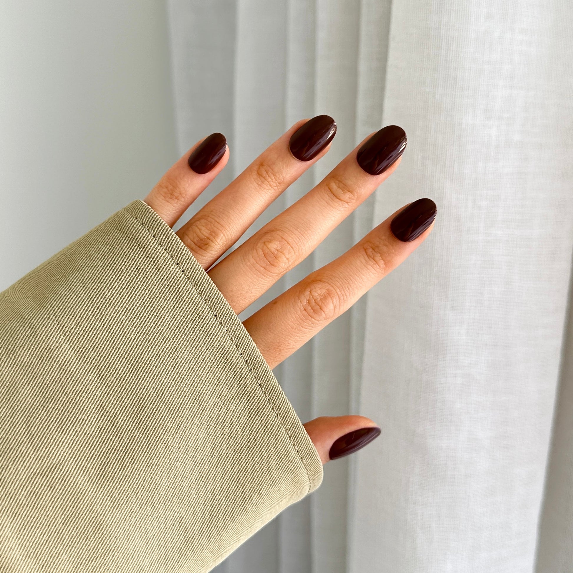 A person's hand with brown oval-shaped press-on nails visible, partially covered by a beige sleeve.