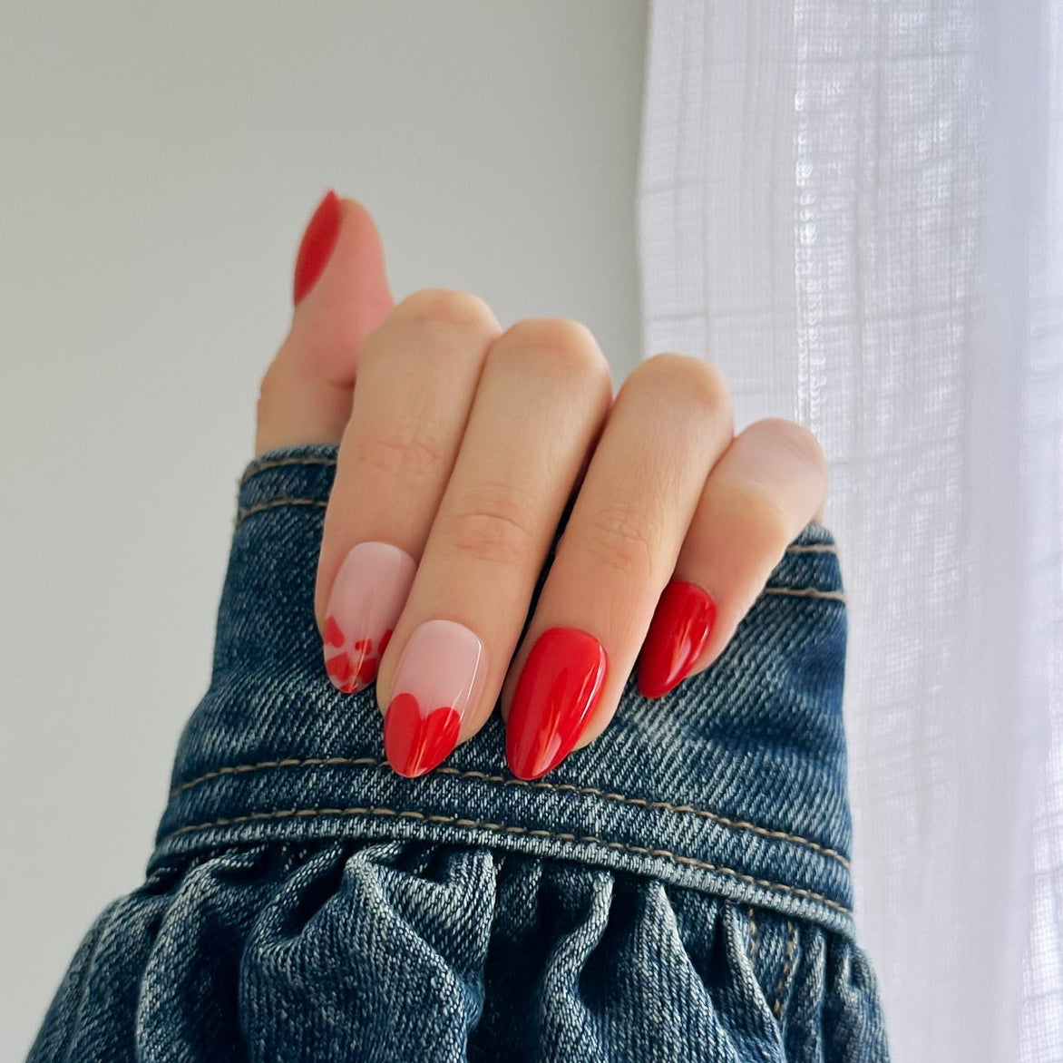 A close-up of a hand with long, glossy red manicured nails resting on the cuff of a blue denim jacket, positioned in front of a white curtain.