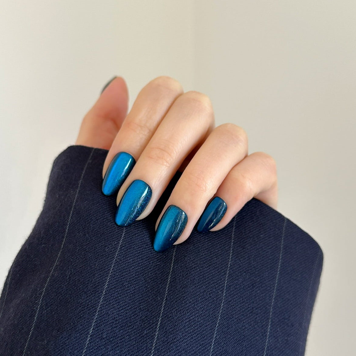 A close-up of a hand with metallic blue manicured nails resting on the cuff of a navy pinstripe blazer against a plain off-white background.
