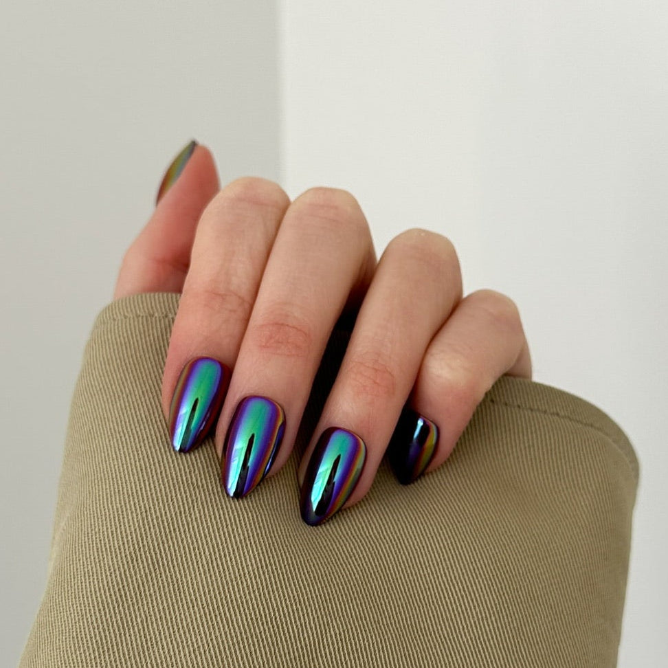 A close-up of a hand with metallic iridescent nail polish in blue, green, and purple tones, resting on a beige sleeve against a plain light gray background.