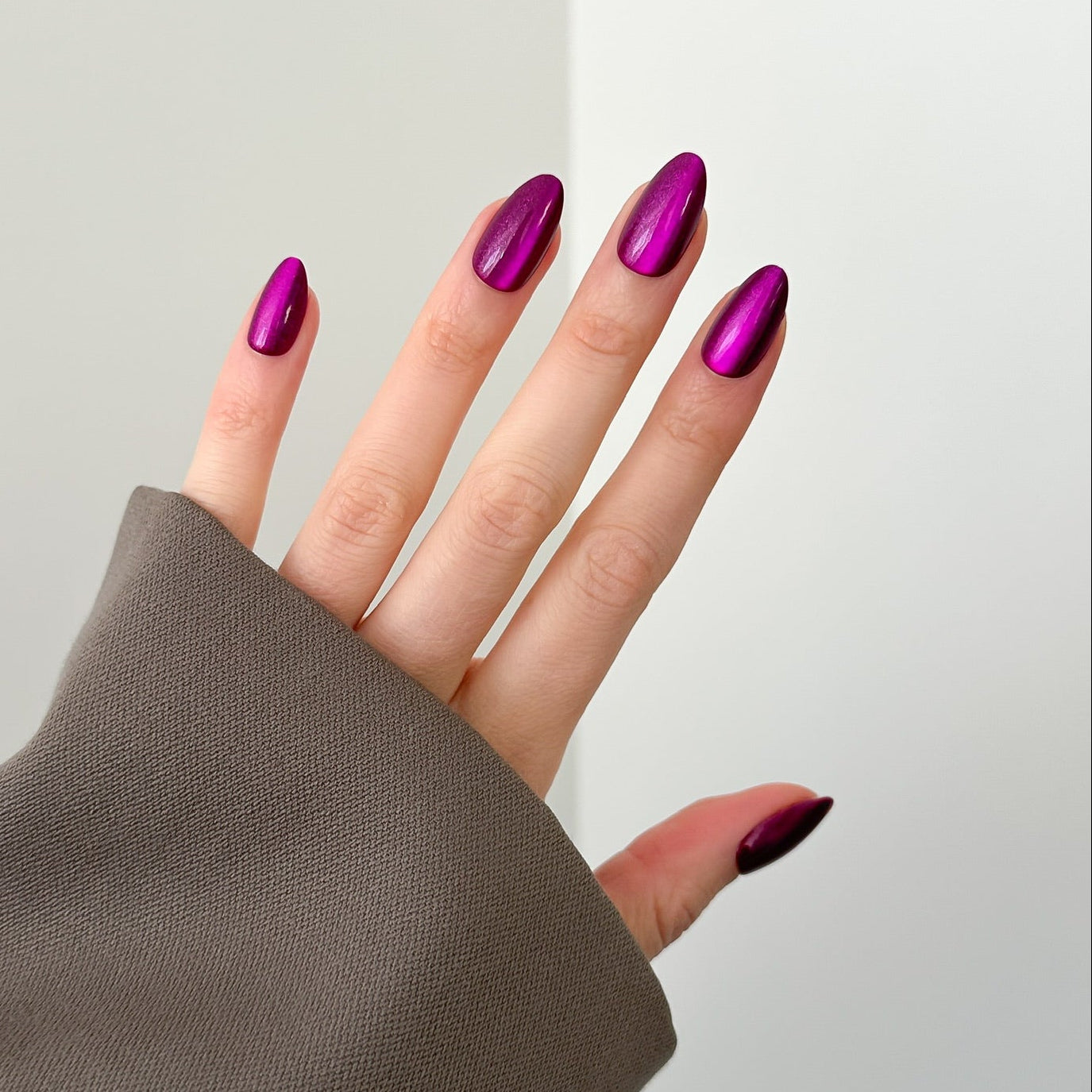 A close-up of a hand with manicured nails painted in a metallic magenta-purple color, wearing a taupe sleeve against a clean white background.