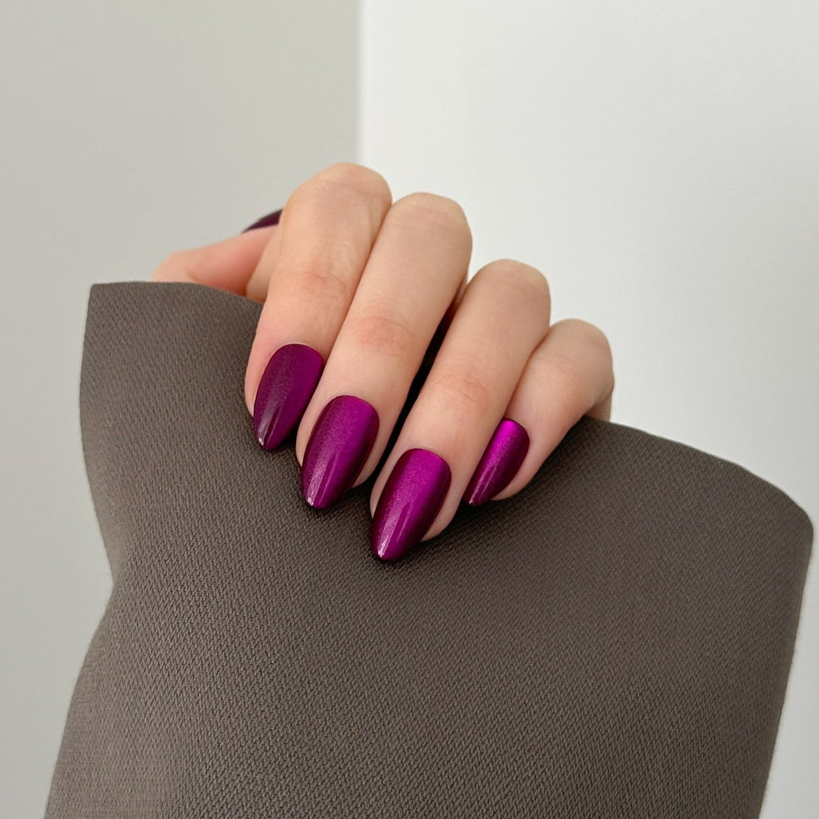 A close-up of a hand with long, almond-shaped nails painted in a glossy deep magenta-purple shade, resting on a textured brown fabric prop against a clean white background.