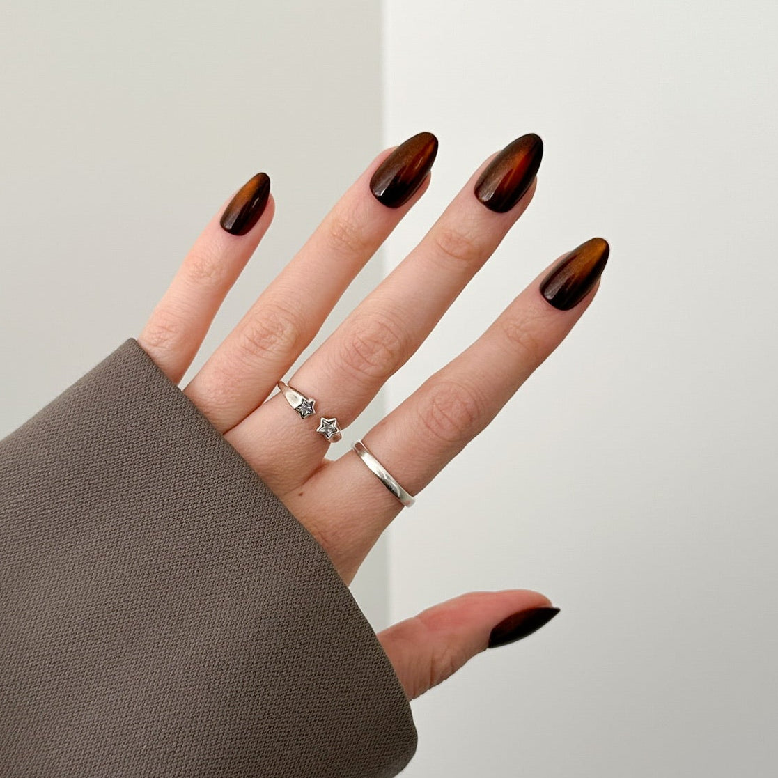 A close-up of a hand with long, glossy dark brown almond-shaped nails, wearing a wide brown cuff sleeve and silver rings, set against a minimalist light gray corner wall.