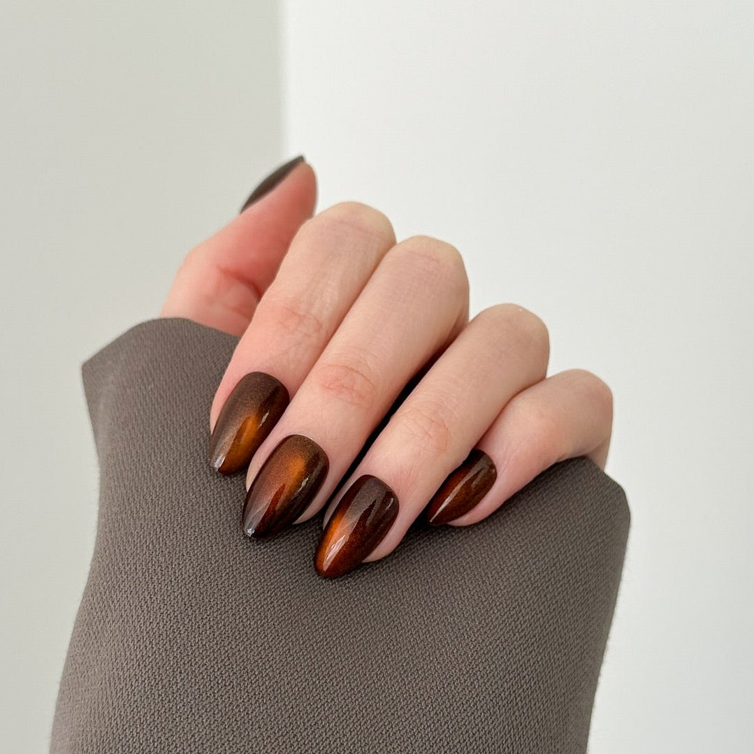 A close-up of a hand with long, glossy brown almond-shaped nails, resting on a textured taupe sleeve against a clean white background.