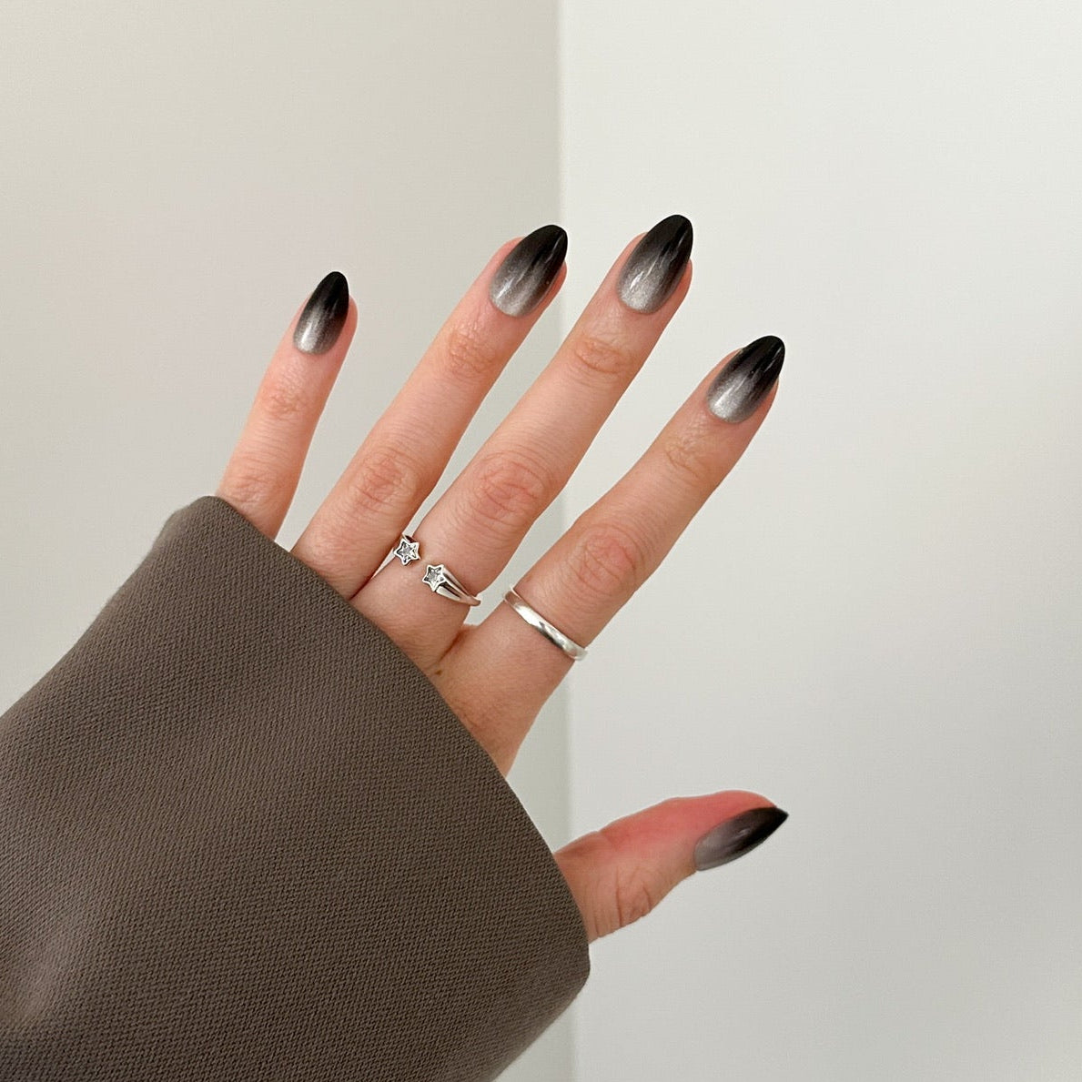 A hand wearing a brown long-sleeve top and silver rings, showcasing almond-shaped nails with a black-to-white ombre gradient, set against a minimalist off-white background.