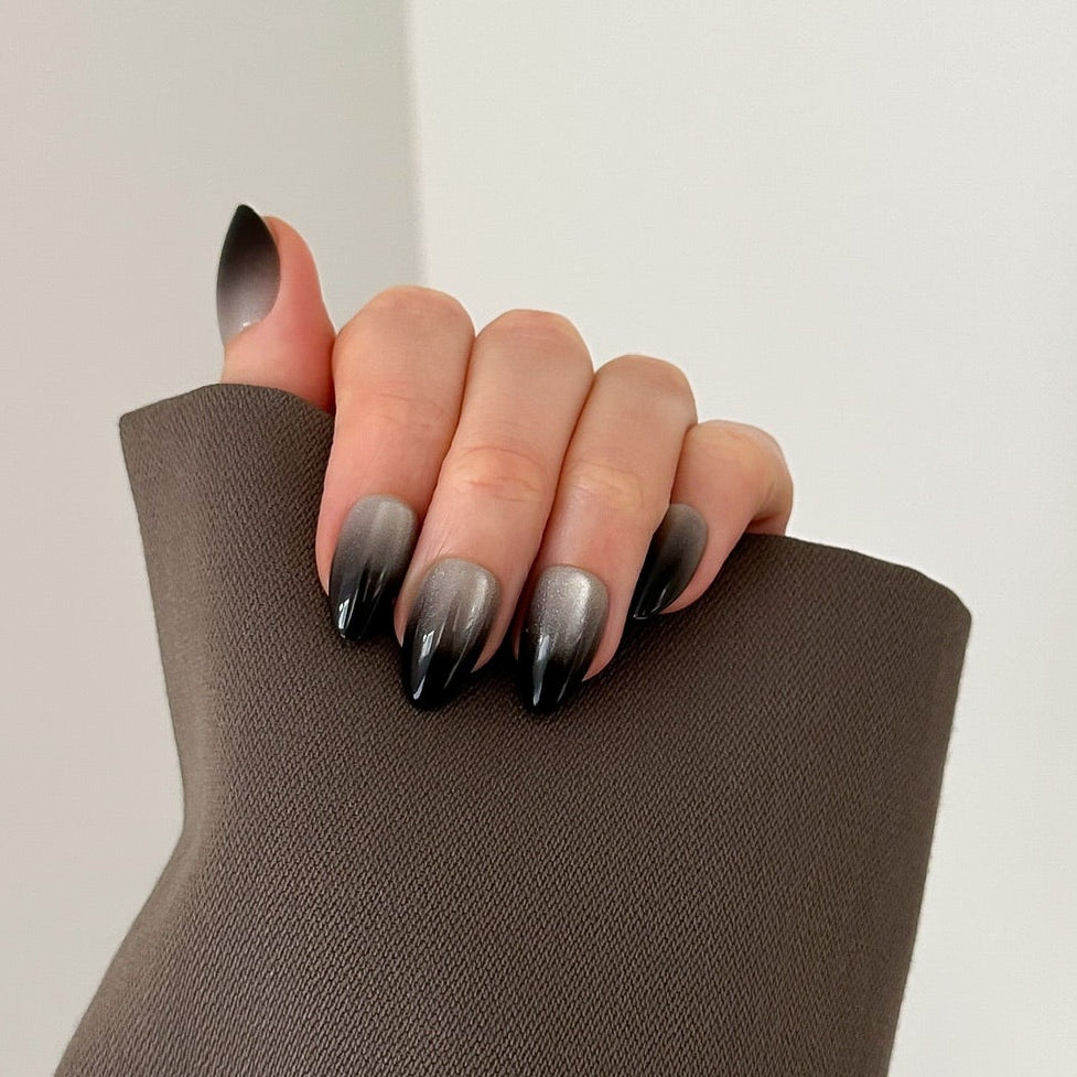 A close-up of a hand with a black-to-gray ombre nail design, resting on the cuff of a taupe-brown sleeve against a plain off-white background.
