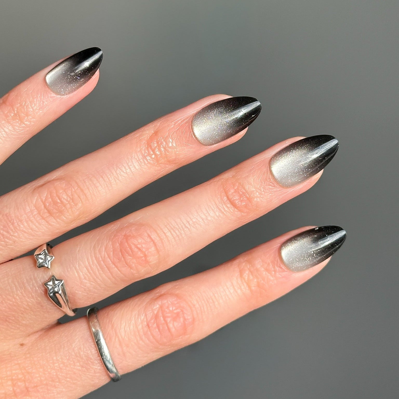 A close-up of a hand with long almond-shaped nails painted in a metallic silver-to-black ombre gradient, wearing silver rings against a solid gray background.