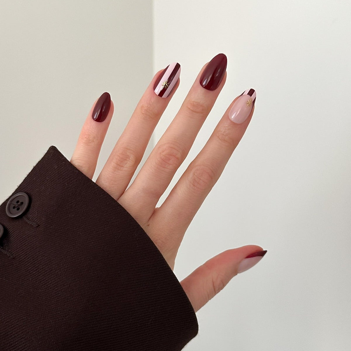 A close-up of a hand with long, almond-shaped nails painted in a dark burgundy and metallic silver manicure, wearing a dark brown blazer sleeve against a clean white background.