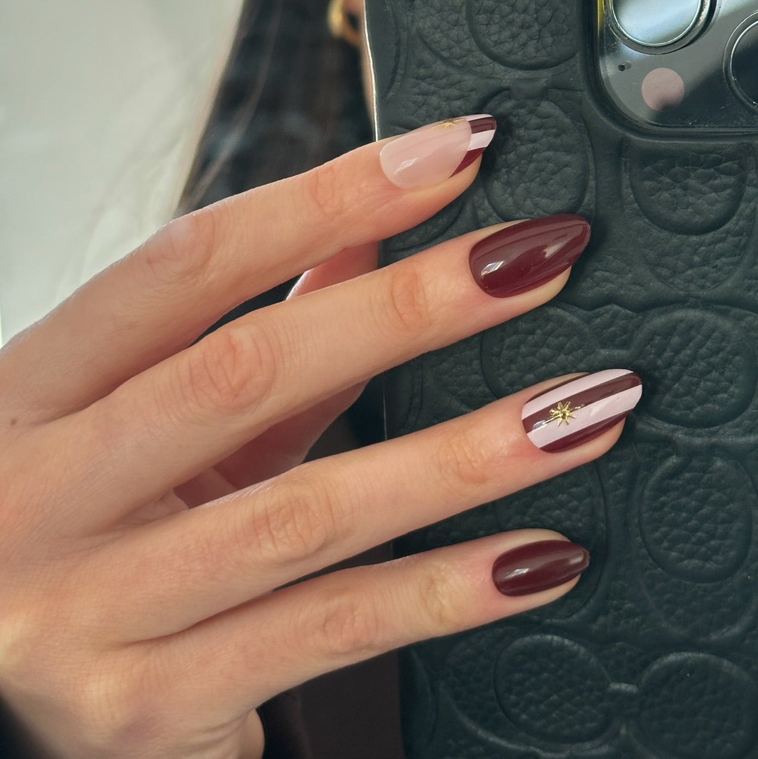 A close-up of a hand with manicured nails, holding a black textured phone case. The nails are styled with a mix of deep burgundy, nude pink, and a white-and-burgundy striped design with a tiny gold star accent.