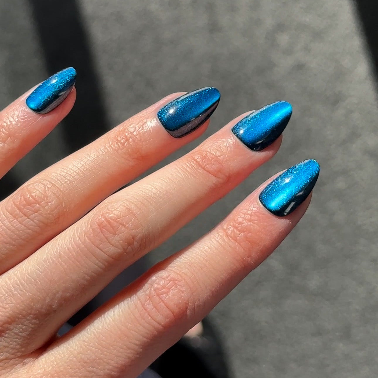 A close-up of a hand with manicured fingernails painted in a metallic, iridescent blue color that shifts with the light, set against a gray textured background.