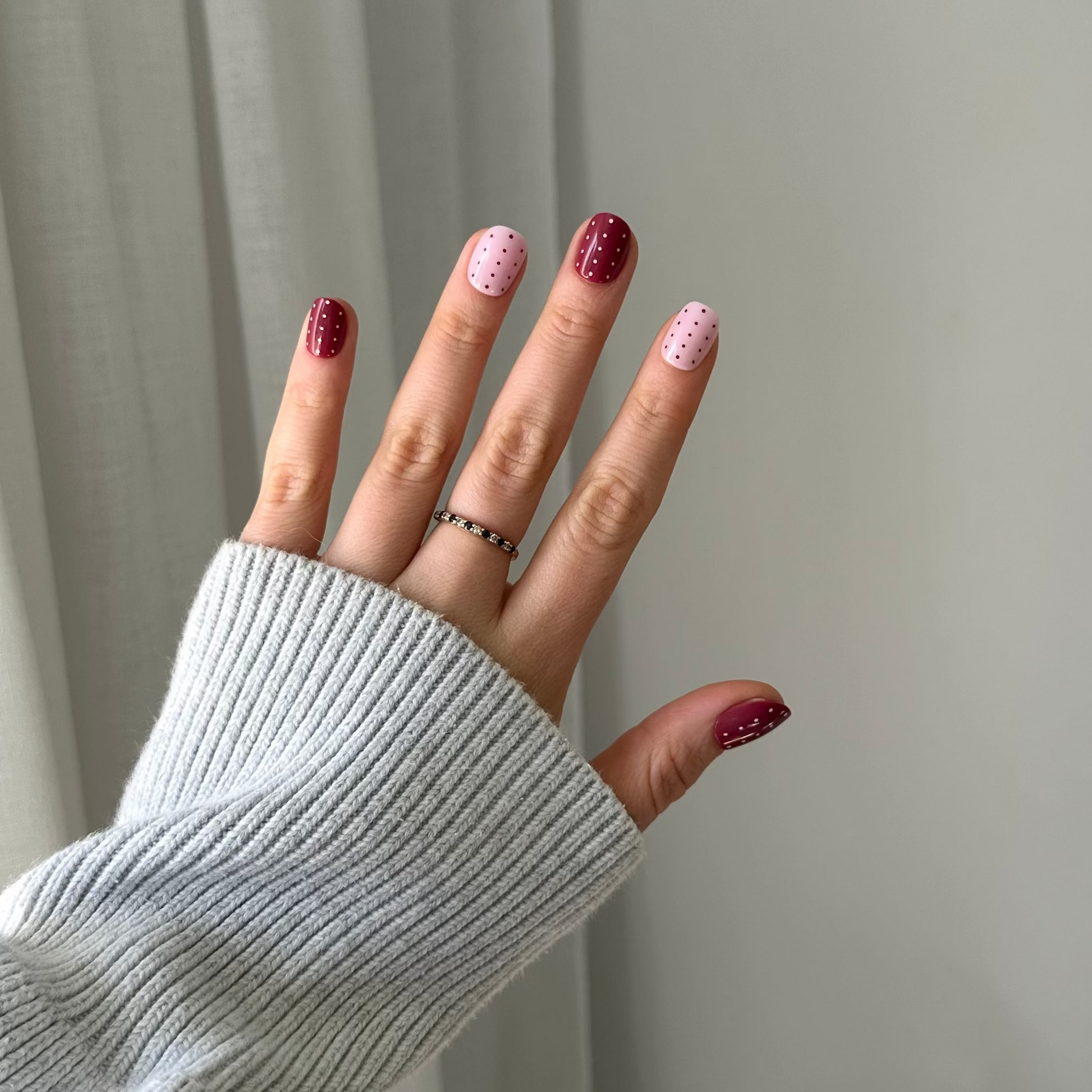 A hand wearing a light gray ribbed knit sweater sleeve, showcasing manicured nails with alternating dark burgundy and light pink glitter polish, along with a thin silver ring.