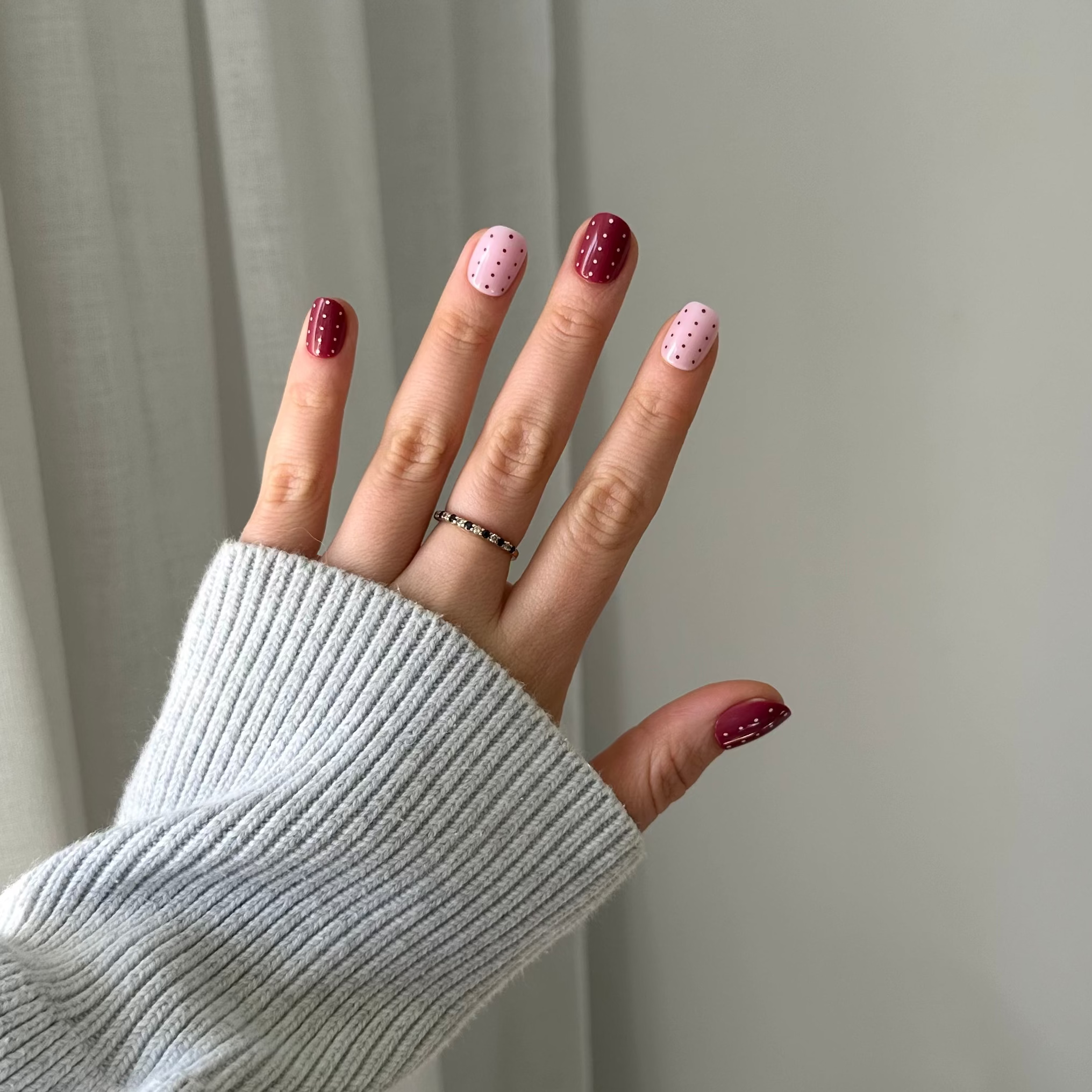 A hand wearing a light gray ribbed knit sweater sleeve, showcasing manicured nails with alternating dark burgundy and light pink glitter polish, along with a thin silver ring.