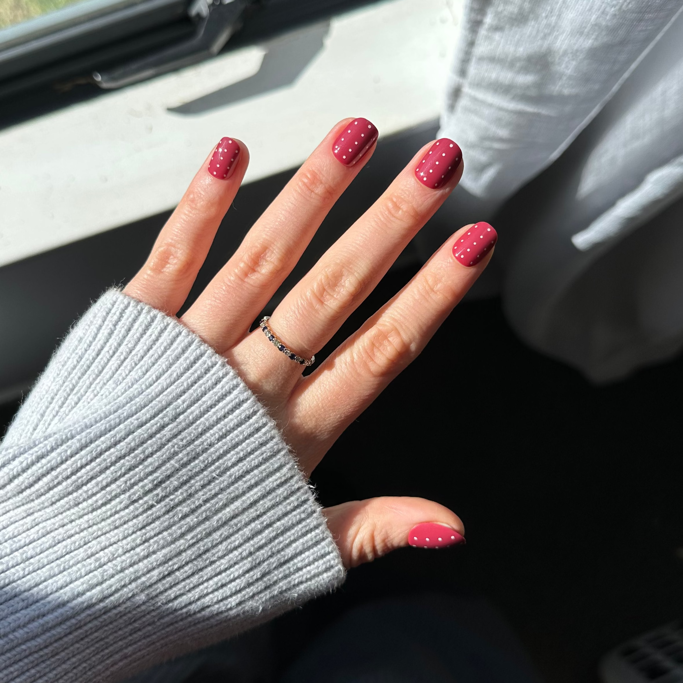 A close-up of a hand with glittery pink nail polish resting near a window, wearing a thin black ring. The person is wearing a light gray ribbed sweater, with a gray curtain and a window showing green grass outside.