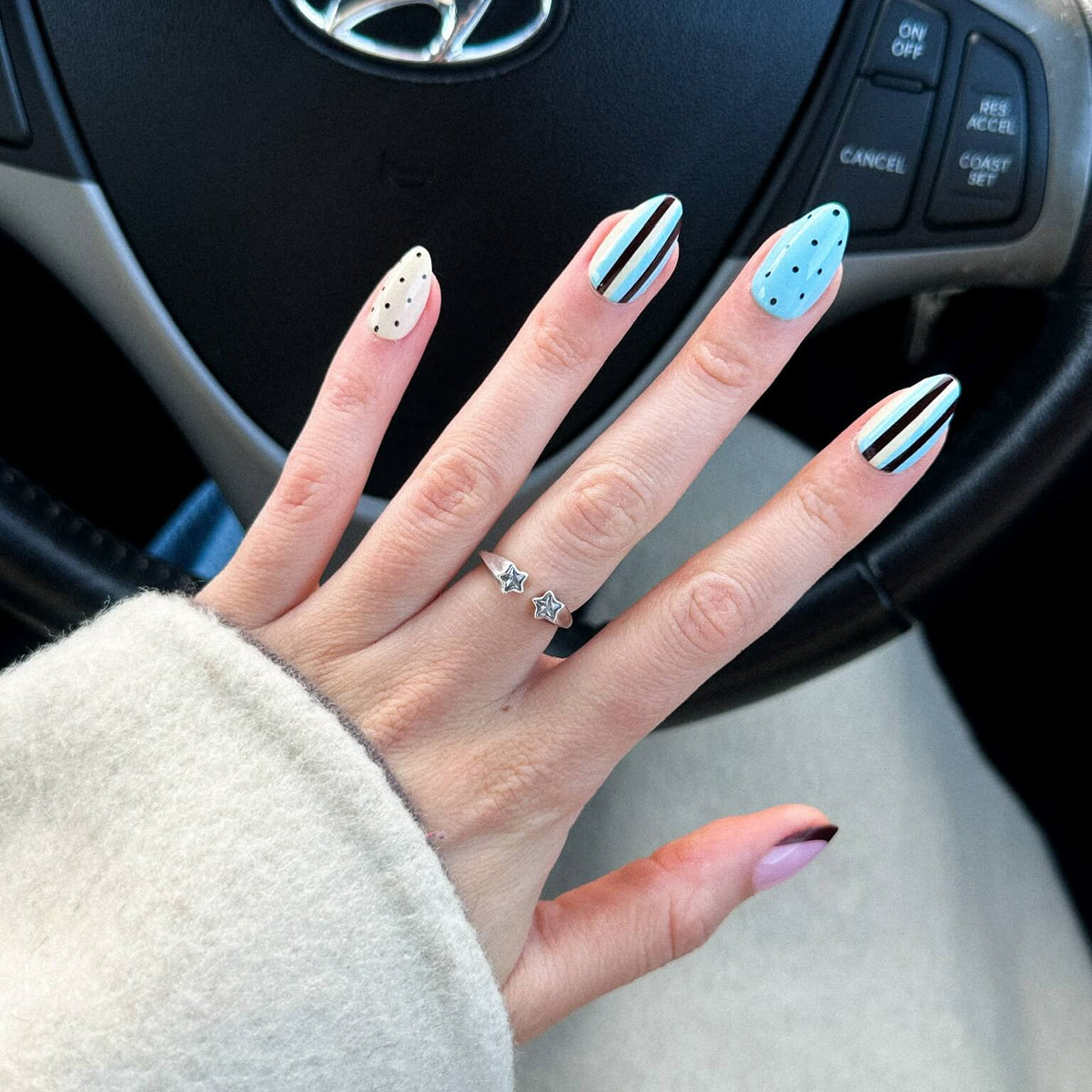 A close-up of a hand with press on nails, resting on a light-colored sweater, with a Hyundai steering wheel in the background. The nails feature a mix of black and white stripes, white speckles, and light blue with black dots.