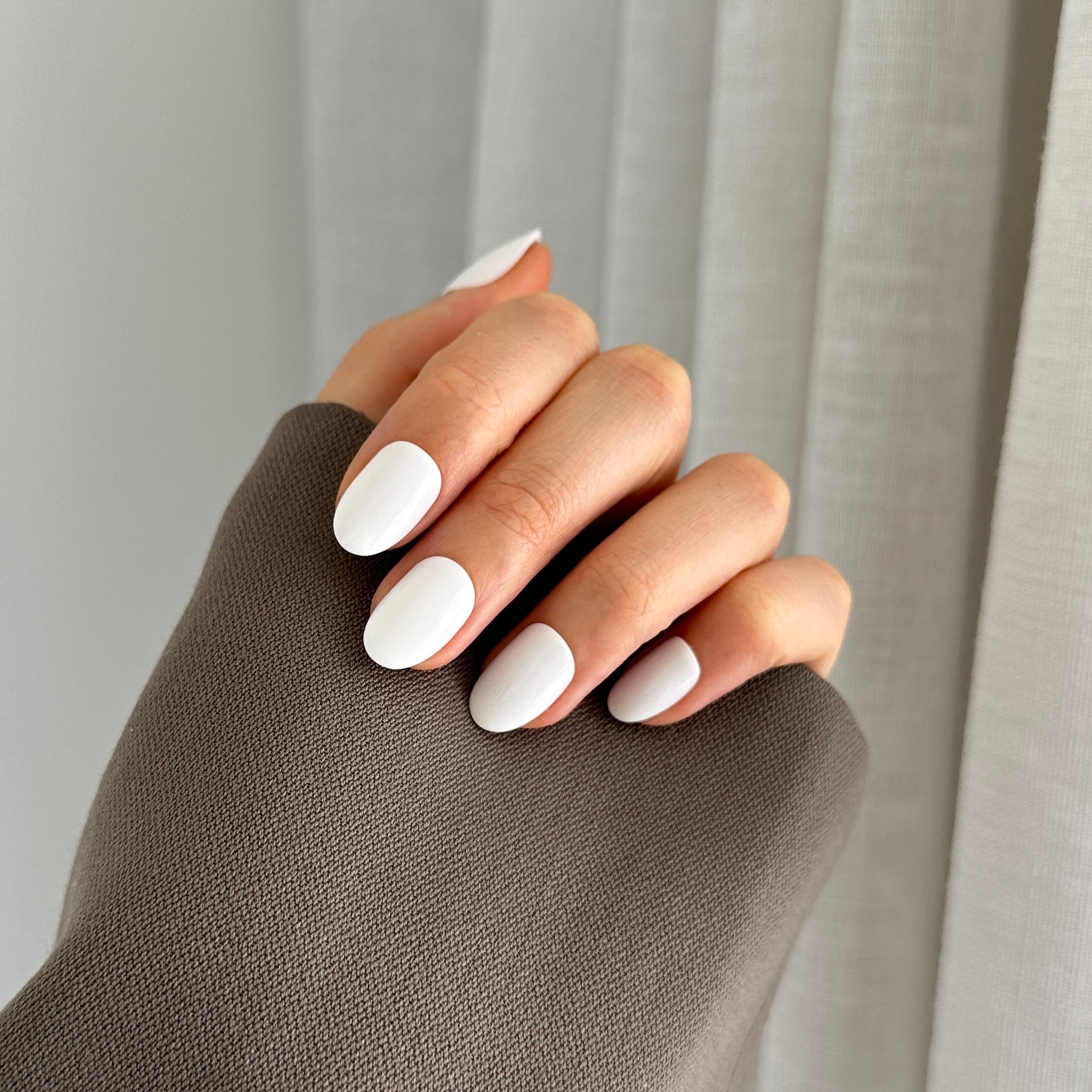 A hand with white oval-shaped press on nails, partially covered by a gray sleeve, against a translucent curtain background.