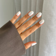 A hand with white oval-shaped press on nails, partially covered by a gray sleeve, against a translucent curtain background.