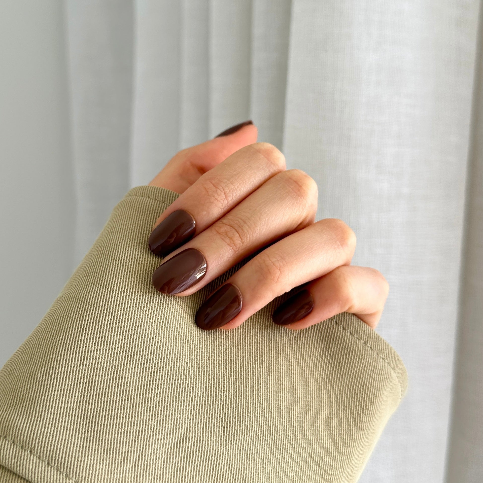 A hand with brown oval-shaped press-on nails, holding a beige object with a curtain in the background.